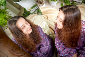 A young woman with long hair wearing a floral robe gazes into a mirror surrounded by plants indoors...