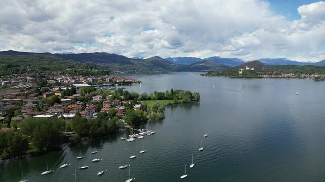 An expansive vista of a tranquil lake, adorned with boats, nestled amidst a picturesque town and rolling hills under a dramatic sky. Aerial view Lake Maggiore, Arona, Italy