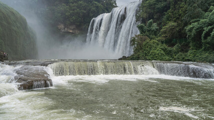 Fototapeta premium Beautiful tropical waterfall. Streams of white water flow down from the cliff. Foam, splashes, haze. Lush green vegetation. In the foreground is a riverbed with rapids. China. Huangguoshu Waterfall. 