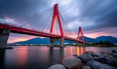 Fototapeta premium Red cable-stayed bridge at sunset over a river, with mountains in the background. Possible use Stock photo for architectural, travel, or nature publications