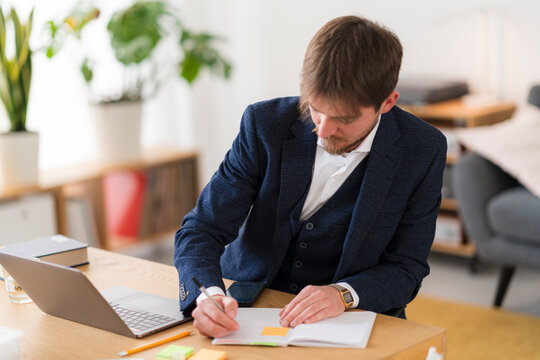 A focused professional in a suit is working at his desk. He is writing in a notebook while using his laptop and phone for work in a bright, modern office.