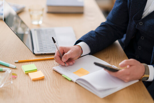 A businessman is at his desk, writing in a notebook while holding his phone. He is wearing a suit jacket and has a laptop nearby.