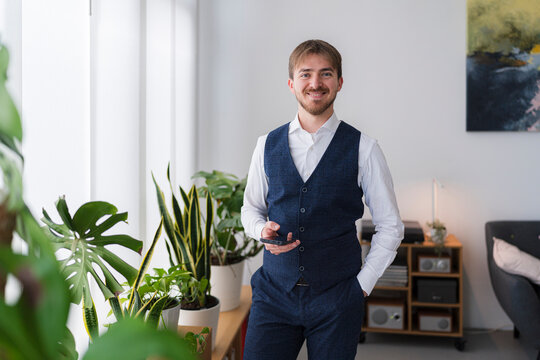 A man in a vest holds a phone in a bright, modern room. He is standing near plants and a window, looking at the camera.