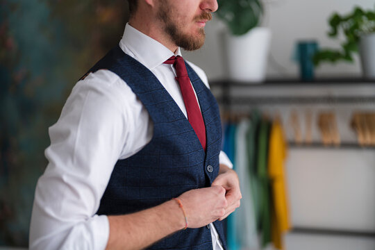 A man fastens his navy vest, wearing a white shirt and red tie. He is getting dressed in front of a rack of colorful clothes.
