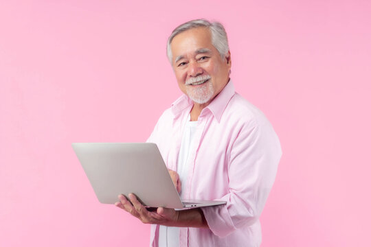Happy Asian senior man using laptop and smiling on pink background, elderly male technology user lifestyle, online communication, retired life, digital literacy in old age, cheerful elder