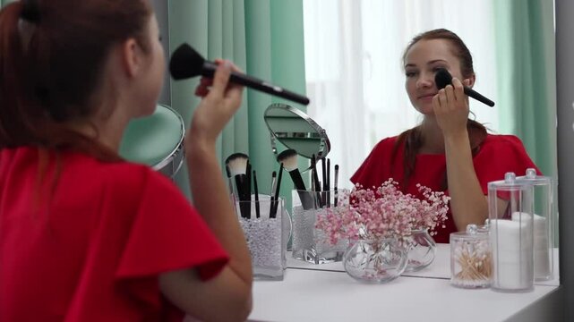 A girl in an evening dress applies powder to her face. Young attractive woman doing makeup behind a dressing table in slow motion.