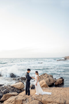 Groom placing wedding ring on bride's finger on rocky coastline.