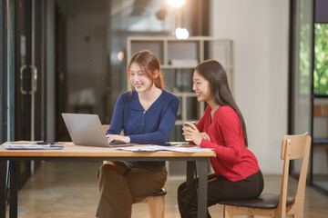 Fototapeta premium Two Asian businesswomen holding graph documents, sitting and talking about work, looking happy while working.