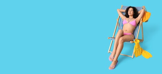 Beautiful young African-American woman in swimsuit relaxing on deck chair against light blue background with space for text