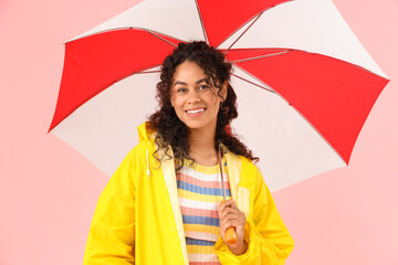 Happy young African-American woman in raincoat with umbrella on pink background
