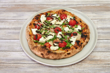 A pizza with cheese and tomatoes on a white plate. The pizza is on a wooden table. The pizza is brown and has a crust