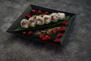 A plate of sushi with a leaf on it is placed on a black plate. The sushi is arranged in a line, with each piece of sushi slightly overlapping the one next to it