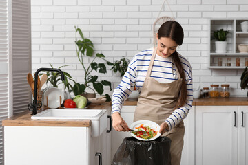 Young woman throwing organic food into trash bin in kitchen. Waste recycling concept