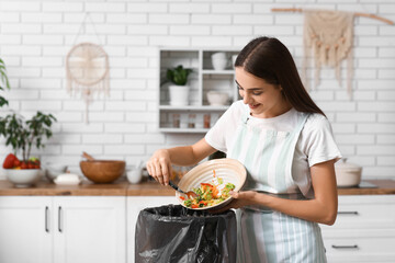 Young woman throwing organic food into trash bin in kitchen. Waste recycling concept