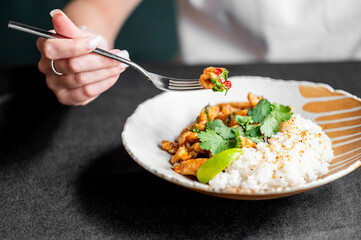 A hand holding a fork with a piece of chicken stir-fry, accompanied by fluffy white rice and garnished with cilantro and lime on a stylish plate.
