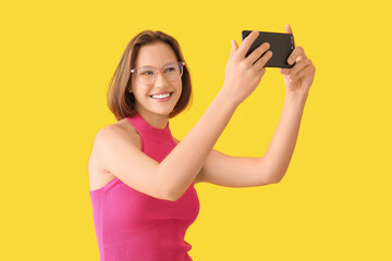 Fototapeta premium Pretty young woman with mobile phone taking selfie on yellow background