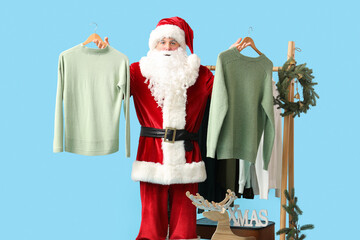 Santa Claus holding hangers with sweaters and Christmas wreath on blue background