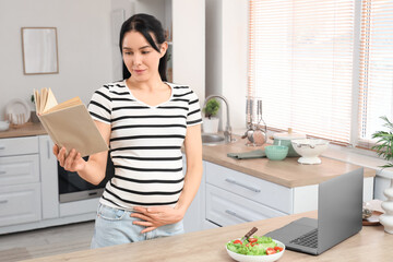 Young pregnant woman reading book in kitchen