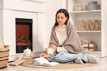Beautiful young happy woman with cup of cacao and plaid reading book near fireplace at home