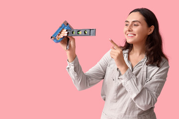 Young female construction worker with bubble level and industrial stapler on pink background. Woman...