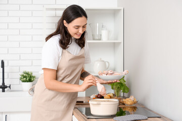 Young housewife with chicken drumsticks cooking soup on stove in kitchen