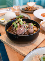 Close up of Thai style traditional pork noodles in a bowl with pork ball and crispy fried garlic in golden color on a table with several foods in scenes, black isolated background.