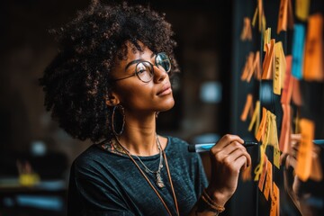 A focused young woman with glasses is brainstorming ideas on a glass board with colorful sticky notes, holding a marker and looking thoughtfully at the notes.