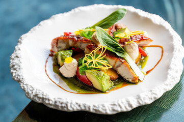 A beautifully plated dish featuring grilled fish with vibrant vegetables, garnished with herbs and decorative elements, served on an artistic white plate.
