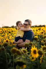 Fototapeta premium Happy laughing father holding baby daughter, standing together in sunflowers meadow. enjoying summer walking together on father's day.