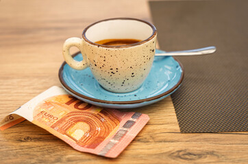 A cup of espresso on a rustic saucer with a 10 euro banknote underneath, placed on a wooden table. Concept of tipping, hospitality, or caf&eacute; culture in Europe.