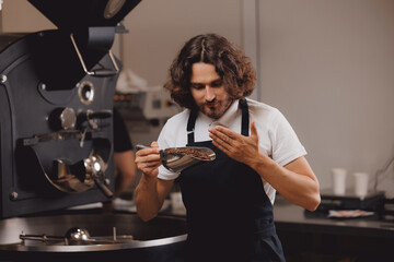 Man barista working with coffee roasting machine on small business factory. Worker testing aroma of fresh hot beans