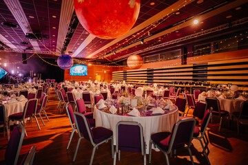Elegant banquet hall decorated with space-themed hanging planets, round tables set for dining, purple chairs, and warm ambient lighting, editorial use..