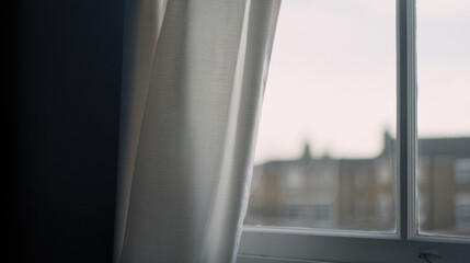 Soft light filters through white curtain hanging by window, with out of focus view of buildings outside, creating calm and peaceful atmosphere