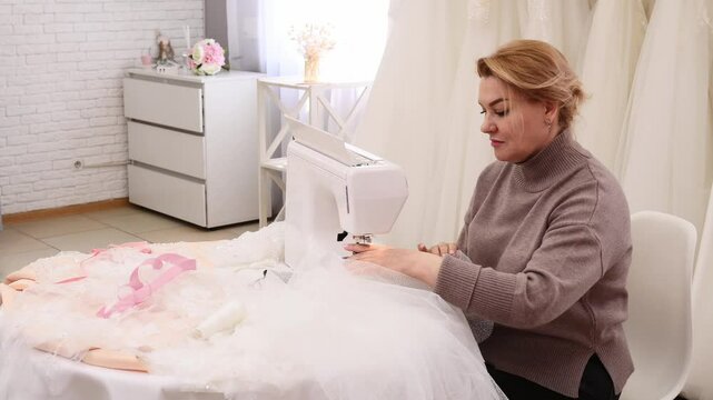 A woman seamstress in a wedding salon sews a bride's dress and veil at a table and with an overlock sewing machine. Handmade, custom tailoring