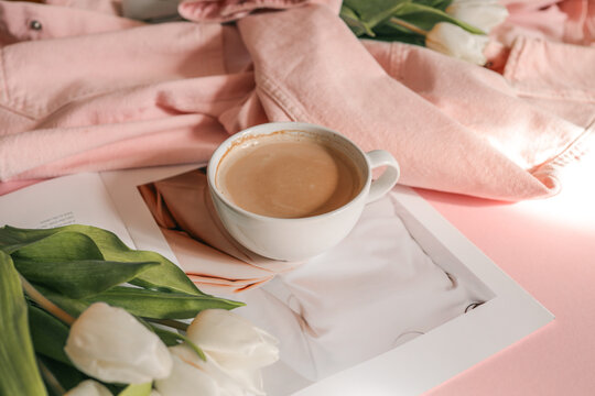 Flat lay with white tulips in a soft pink denim jacket, a fashion magazine, and a cup of coffee on a pastel pink background - cozy, stylish morning scene with feminine spring mood