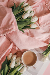Flat lay with white tulips in a soft pink denim jacket, a fashion magazine, and a cup of coffee on a pastel pink background — cozy, stylish morning scene with feminine spring mood