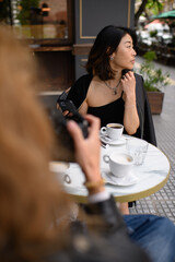 Asian woman sitting at an outdoor café table, holding a smartphone and enjoying her coffee. Relaxed moment of modern lifestyle, street atmosphere, and digital connection.