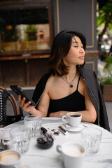 Asian woman sitting at an outdoor café table, holding a smartphone and enjoying her coffee. Relaxed moment of modern lifestyle, street atmosphere, and digital connection.
