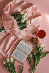 White tulips, open Bible, cup of tea, and candle on a pink background with a soft pink denim jacket...