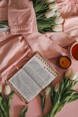 White tulips, open Bible, cup of tea, and candle on a pink background with a soft pink denim jacket...