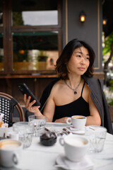 Asian woman sitting at an outdoor café table, holding a smartphone and enjoying her coffee. Relaxed moment of modern lifestyle, street atmosphere, and digital connection.