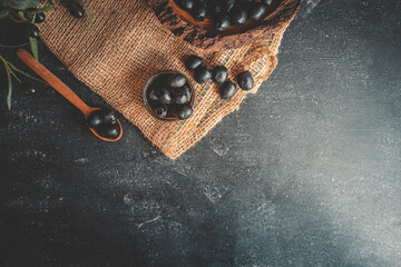 Top view of rustic composition with black olives in a wooden bowl, a wooden spoon, a glass bottle with cork, and burlap on a dark background. Warm earthy food scene