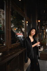 Asian woman standing alone by a café bar counter on a city street, enjoying her morning coffee and quietly observing the urban scenery before starting her workday.