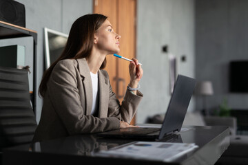 Young woman sitting at office desk, holding a pen to her chin in deep thought. laptop is open in front of her