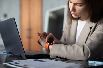 Young female professional is sitting at a desk in an office using smartwatch on her wrist.