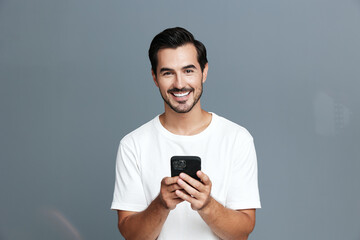 Smiling man in casual white t-shirt holding smartphone while looking at the camera with a joyful expression against a plain grey background. People lifestyle concept.