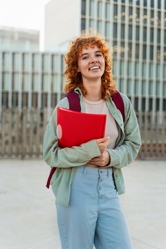 University student portrait of red-haired girl looking at the camera with a folder and backpack smiling 