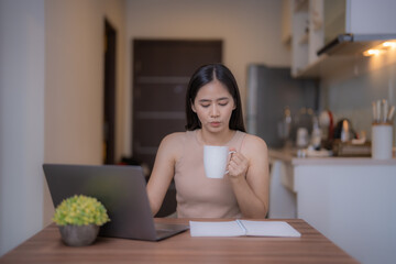 Young Asian woman enjoying coffee while working from her home office, using a laptop and notebook...