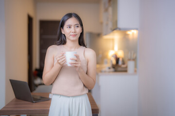 Serene young Asian woman enjoys a quiet moment with a cup of coffee in her modern kitchen, her gaze...