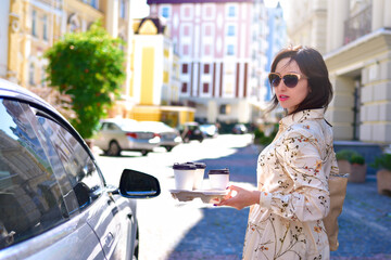 A smiling woman wearing sunglasses is holding a coffee in her hands near a car against the backdrop of a city